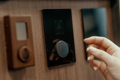 A close-up view of a person's hand adjusting a digital thermostat on a wooden wall. The thermostat has a circular knob and a small display showing the temperature. Next to it, there is a smaller device with a teardrop-shaped button.