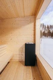 A cozy wooden sauna with light-colored wood paneling covers the ceiling, walls, and floor. A modern black stove is placed near a large window offering a view of snowy trees outside.