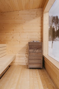 A cozy sauna with light wooden walls, floor, and ceiling, complemented by a large window pane. A wooden bench lines one side, and a wooden box with stones on top is positioned against the wall.