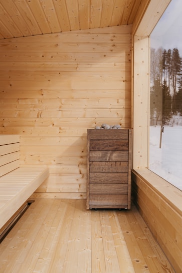 A cozy sauna with light wooden walls, floor, and ceiling, complemented by a large window pane. A wooden bench lines one side, and a wooden box with stones on top is positioned against the wall.