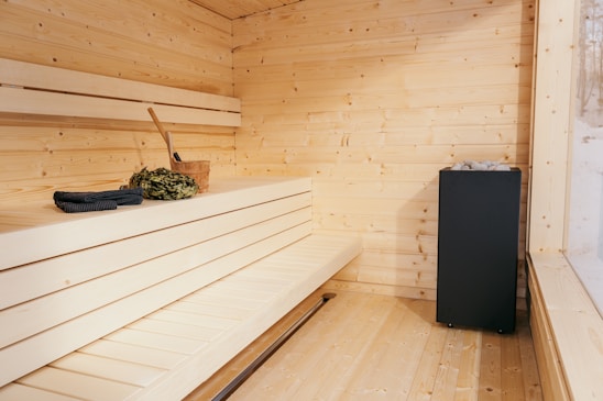 A wooden sauna room with benches made of light-colored wood. A bucket with a ladle and a bundle of leaves are placed on the bench. A black heater with stones is located in the corner, and a large window provides natural light.