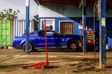 A blue car is parked inside a mechanic's workshop. The car is raised on a jack with its front wheel removed, indicating ongoing repair or maintenance work. Various tools and equipment are visible around the car, including a red hydraulic jack in the foreground and several mechanics' tools hanging from the ceiling. The workshop is partially open, with sunlight casting shadows on the concrete floor and blue corrugated metal walls.