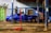 A blue car is parked inside a mechanic's workshop. The car is raised on a jack with its front wheel removed, indicating ongoing repair or maintenance work. Various tools and equipment are visible around the car, including a red hydraulic jack in the foreground and several mechanics' tools hanging from the ceiling. The workshop is partially open, with sunlight casting shadows on the concrete floor and blue corrugated metal walls.