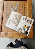 A book with colorful illustrations is open on a wooden table. The table has a rustic appearance with visible text and patterns. Below the table, a person wearing black and white sneakers and a striped skirt is partially visible.