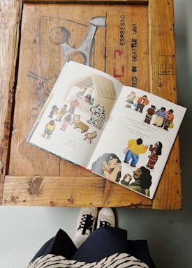 A book with colorful illustrations is open on a wooden table. The table has a rustic appearance with visible text and patterns. Below the table, a person wearing black and white sneakers and a striped skirt is partially visible.
