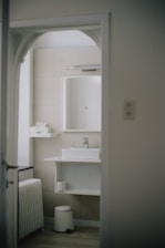 A modern bathroom with minimalist design features a white sink on a countertop with a mirror above. The walls are tiled in a neutral beige color. A small shelf holds toiletries, and a stool sits beneath the sink. The bathroom is viewed through an arched doorway.