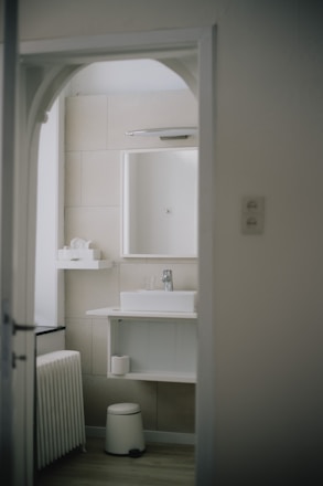 A modern bathroom with minimalist design features a white sink on a countertop with a mirror above. The walls are tiled in a neutral beige color. A small shelf holds toiletries, and a stool sits beneath the sink. The bathroom is viewed through an arched doorway.