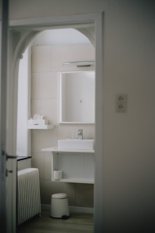 A modern bathroom with minimalist design features a white sink on a countertop with a mirror above. The walls are tiled in a neutral beige color. A small shelf holds toiletries, and a stool sits beneath the sink. The bathroom is viewed through an arched doorway.