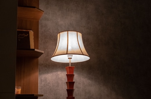 A warmly lit table lamp with a classic fabric shade stands against a textured gray wall. To the left, a wooden shelf holds a few books.