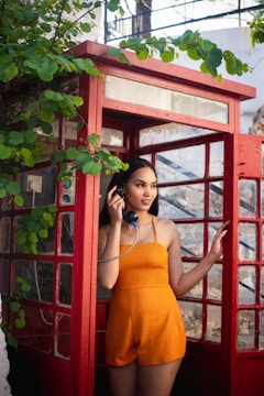 A woman stands in a vintage red telephone booth, holding a phone to her ear. She is wearing an orange sleeveless outfit and is surrounded by green foliage. The setting appears to be outdoors with natural lighting.
