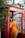 A woman stands in a vintage red telephone booth, holding a phone to her ear. She is wearing an orange sleeveless outfit and is surrounded by green foliage. The setting appears to be outdoors with natural lighting.