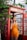 A woman stands in a vintage red telephone booth, holding a phone to her ear. She is wearing an orange sleeveless outfit and is surrounded by green foliage. The setting appears to be outdoors with natural lighting.