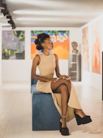 A person sits elegantly on a blue bench in a well-lit art gallery, wearing a beige sleeveless dress and black platform shoes. The walls display various colorful abstract paintings and a sculpture bust, creating a sophisticated and artistic atmosphere.