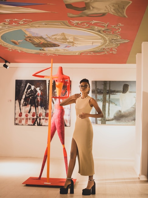 A stylish woman in a beige dress and high heels stands confidently in an art gallery. Behind her is a tall, abstract red sculpture and several large artworks. The room is well-lit with a vibrant painted ceiling featuring mythical and nautical themes.