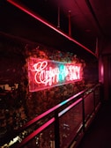 Neon sign in bright red and blue colors hangs prominently on a wall decorated with lush green ivy. The sign emits a warm glow that contrasts with the dark surroundings, creating a vibrant effect against the metal railing nearby.