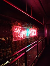 Neon sign in bright red and blue colors hangs prominently on a wall decorated with lush green ivy. The sign emits a warm glow that contrasts with the dark surroundings, creating a vibrant effect against the metal railing nearby.