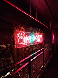 Neon sign in bright red and blue colors hangs prominently on a wall decorated with lush green ivy. The sign emits a warm glow that contrasts with the dark surroundings, creating a vibrant effect against the metal railing nearby.