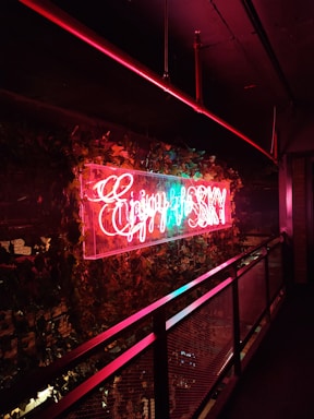 Neon sign in bright red and blue colors hangs prominently on a wall decorated with lush green ivy. The sign emits a warm glow that contrasts with the dark surroundings, creating a vibrant effect against the metal railing nearby.