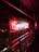 Neon sign in bright red and blue colors hangs prominently on a wall decorated with lush green ivy. The sign emits a warm glow that contrasts with the dark surroundings, creating a vibrant effect against the metal railing nearby.
