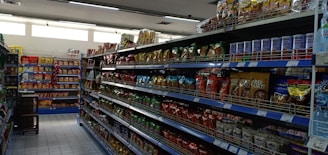 A supermarket aisle filled with various packaged snacks and food items. Shelves on both sides are stocked with colorful bags and packets, including chips and canned goods. The lighting is bright, and the floor is tiled.
