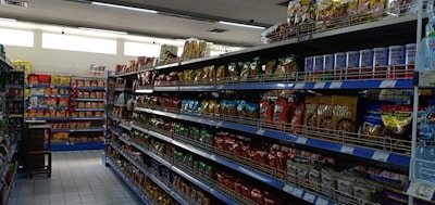 A supermarket aisle filled with various packaged snacks and food items. Shelves on both sides are stocked with colorful bags and packets, including chips and canned goods. The lighting is bright, and the floor is tiled.