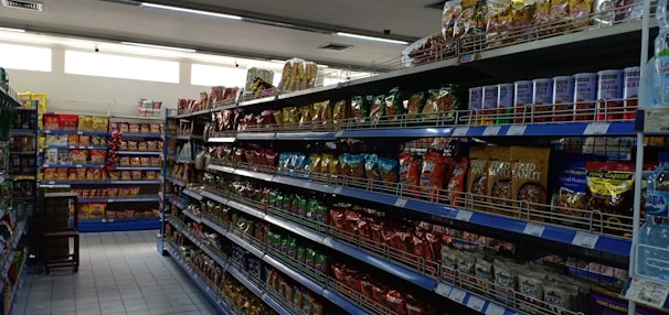 A supermarket aisle filled with various packaged snacks and food items. Shelves on both sides are stocked with colorful bags and packets, including chips and canned goods. The lighting is bright, and the floor is tiled.