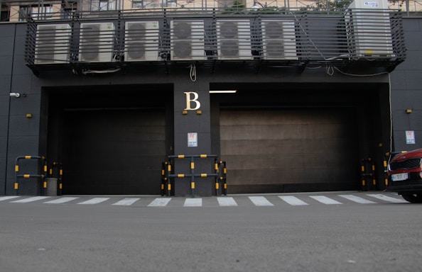 A large industrial garage door is situated in a dark gray building facade, marked with the letter 'B' above it. Air conditioning units are visible on a platform above the door. Yellow and black striped barriers are positioned on either side of the entrance. A zebra crossing is seen in front of the building, with the front of a red car partially in view on the right.