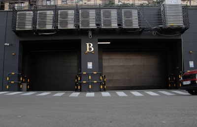 A large industrial garage door is situated in a dark gray building facade, marked with the letter 'B' above it. Air conditioning units are visible on a platform above the door. Yellow and black striped barriers are positioned on either side of the entrance. A zebra crossing is seen in front of the building, with the front of a red car partially in view on the right.