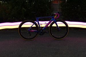 A vibrant road bike with a blue frame is positioned on a dark asphalt surface. The bike's frame has the brand name embossed in pink letters. Behind the bike, a mesmerizing trail of light in pink and yellow hues creates a dynamic visual effect, suggesting motion or light painting. The background includes lush greenery and a fence partially visible.