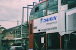 A two-story bus with advertising on its side is parked in an urban environment. The advertisement displays the brand 'DAIKIN' and some text in a foreign script. There is construction scaffolding in the background, along with residential buildings and a parked car.