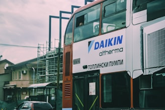 A two-story bus with advertising on its side is parked in an urban environment. The advertisement displays the brand 'DAIKIN' and some text in a foreign script. There is construction scaffolding in the background, along with residential buildings and a parked car.