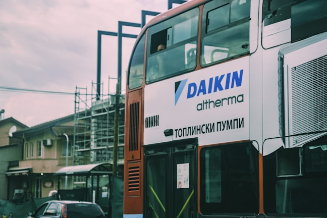 A two-story bus with advertising on its side is parked in an urban environment. The advertisement displays the brand 'DAIKIN' and some text in a foreign script. There is construction scaffolding in the background, along with residential buildings and a parked car.