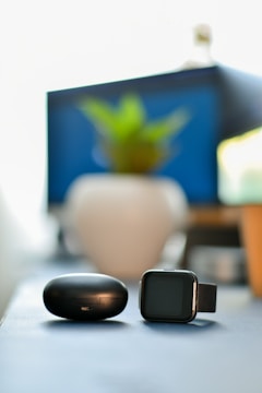A black smartwatch and a black round charging case are placed on a flat surface. A blurred background features a potted plant with green leaves, a framed blue object, and soft natural lighting.