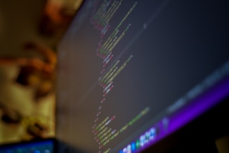 A close-up view of a computer monitor displaying lines of code with a blurred background. The text on the screen is code, likely from a programming language, with syntax highlighting showing different colors for keywords, variables, and strings.