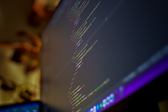 A close-up view of a computer monitor displaying lines of code with a blurred background. The text on the screen is code, likely from a programming language, with syntax highlighting showing different colors for keywords, variables, and strings.