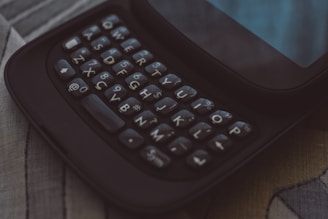 A close-up view of a mobile device featuring a physical QWERTY keyboard. The keys are prominently displayed, with white characters on a dark background. The screen above the keyboard is partially visible, and the scene is set on a textured surface.
