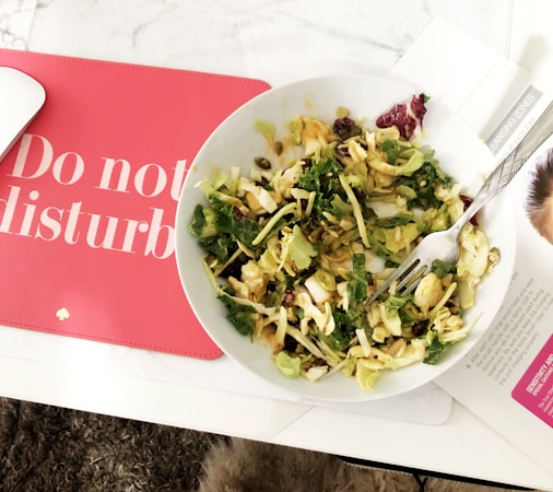 A bowl of salad containing green leafy vegetables, possibly kale and cabbage, placed on a white table. Beside it, a red mat with the words 'Do not disturb' is partially visible. There is a silver fork in the bowl, and part of a magazine or document with text can be seen underneath the bowl.