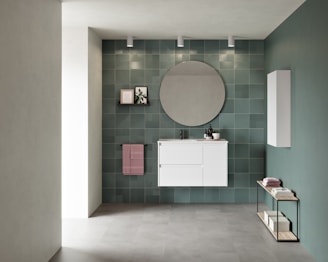 A modern bathroom interior featuring a white vanity with drawers mounted on a wall of green tiles. Above the vanity, a large circular mirror is flanked by three ceiling spotlights. To the left, a small shelf holds a framed picture and a plant, while a pink towel hangs below it. An additional white cabinet is installed on the adjacent wall. A minimalist console table with books and towels is placed on the floor, emphasizing a contemporary aesthetic.