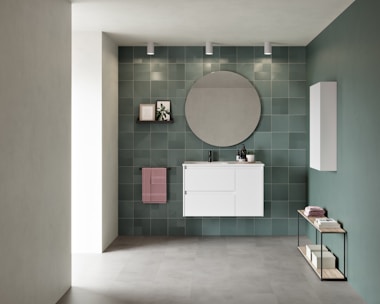A modern bathroom interior featuring a white vanity with drawers mounted on a wall of green tiles. Above the vanity, a large circular mirror is flanked by three ceiling spotlights. To the left, a small shelf holds a framed picture and a plant, while a pink towel hangs below it. An additional white cabinet is installed on the adjacent wall. A minimalist console table with books and towels is placed on the floor, emphasizing a contemporary aesthetic.
