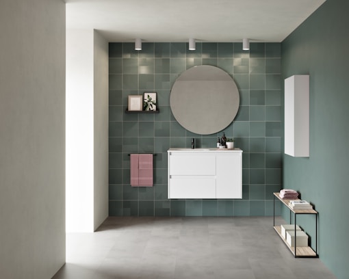 A modern bathroom interior featuring a white vanity with drawers mounted on a wall of green tiles. Above the vanity, a large circular mirror is flanked by three ceiling spotlights. To the left, a small shelf holds a framed picture and a plant, while a pink towel hangs below it. An additional white cabinet is installed on the adjacent wall. A minimalist console table with books and towels is placed on the floor, emphasizing a contemporary aesthetic.