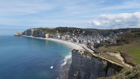 A coastal landscape featuring striking white cliffs that stretch along a clear blue sea. A quaint town with a collection of small buildings lines the shoreline. The sky is partly cloudy, adding depth to the scene. The foreground includes grassy areas and pathways where several people can be seen strolling.