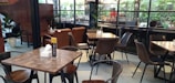 The image features a modern cafe interior with several wooden tables and chairs arranged neatly. Large glass windows with dark frames offer a view of lush green plants outside. The space is well-lit and decorated with a minimalistic style.