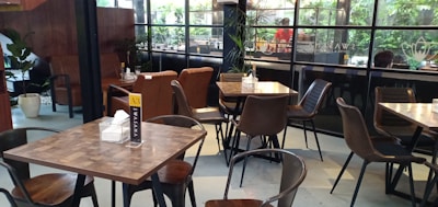 The image features a modern cafe interior with several wooden tables and chairs arranged neatly. Large glass windows with dark frames offer a view of lush green plants outside. The space is well-lit and decorated with a minimalistic style.