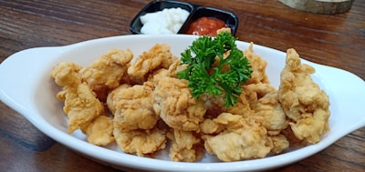 A white oval dish filled with crispy fried chicken pieces garnished with fresh green parsley. In the background, there are two small black bowls containing diced white onions and red dipping sauce, all placed on a wooden table.