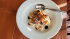 A white plate contains a serving of oatmeal topped with assorted dried fruits and nuts. A metal spoon is resting on the side of the plate, and the setup is placed on a wooden table with soft natural lighting creating shadows.