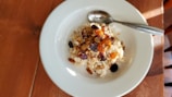 A white plate contains a serving of oatmeal topped with assorted dried fruits and nuts. A metal spoon is resting on the side of the plate, and the setup is placed on a wooden table with soft natural lighting creating shadows.