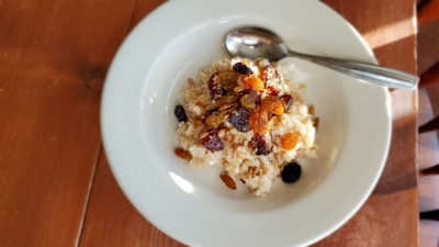 A white plate contains a serving of oatmeal topped with assorted dried fruits and nuts. A metal spoon is resting on the side of the plate, and the setup is placed on a wooden table with soft natural lighting creating shadows.