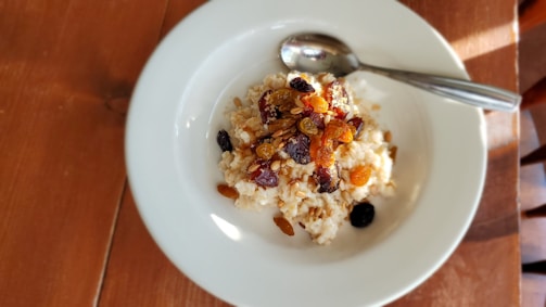 A white plate contains a serving of oatmeal topped with assorted dried fruits and nuts. A metal spoon is resting on the side of the plate, and the setup is placed on a wooden table with soft natural lighting creating shadows.