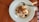 A white plate contains a serving of oatmeal topped with assorted dried fruits and nuts. A metal spoon is resting on the side of the plate, and the setup is placed on a wooden table with soft natural lighting creating shadows.