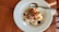 A white plate contains a serving of oatmeal topped with assorted dried fruits and nuts. A metal spoon is resting on the side of the plate, and the setup is placed on a wooden table with soft natural lighting creating shadows.